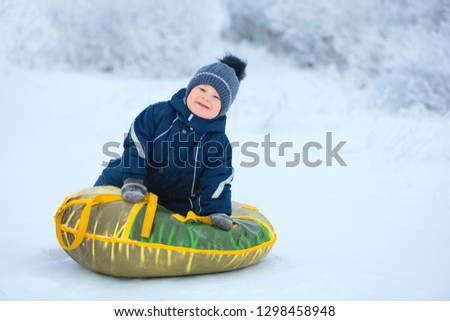 The portrait of caucasian child for a walk with a color inflatable sled in winter. The male child, 3 years old, is wearing the blue jumpsuit, the warm cap and the grey mittens is outdoors.