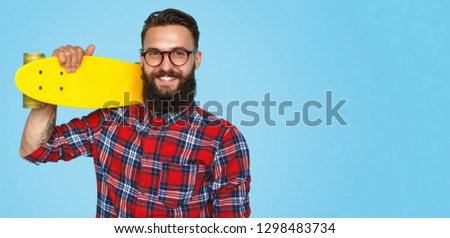 Happy bearded hipster in glasses and plaid shirt holding bright skateboard on blue backdrop