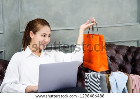 Beautiful Asian woman is smiling.Lady work with laptops on the sofa in the room in the morning.Orange shopping bag in his hand.Girl in white shirt.