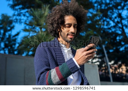 Portrait of a young handsome African man using his smartphone with smile while sitting on a bench outdoors in sunny day