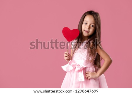 portrait of a cute little girl in a pink dress holding a paper heart on a pink background. St. Valentine's Day