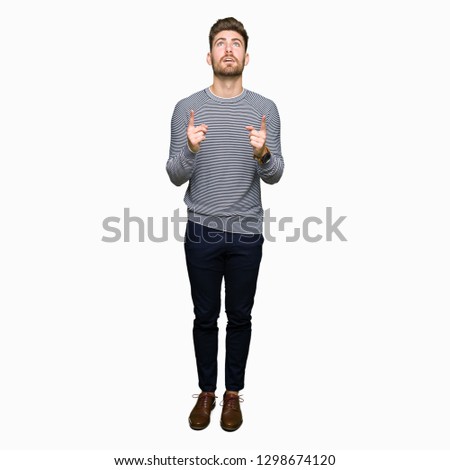 Young handsome man wearing stripes sweater amazed and surprised looking up and pointing with fingers and raised arms.