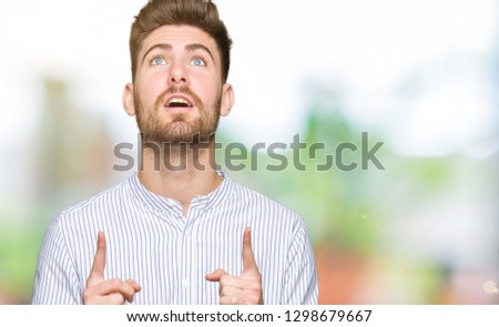 Young handsome man amazed and surprised looking up and pointing with fingers and raised arms.