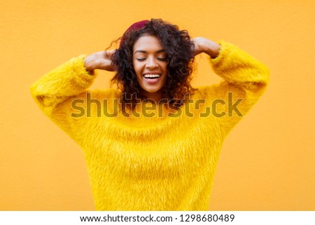 Blissful  black  smiling woman  with clothes eyes  playing with her curly hair on yellow background in cute fluffy  sweater and   Burgundy beret. 