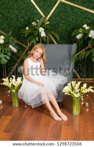 Little cute girl with blond hair in a white dress and white flowers, lilies and orchids on a green background
