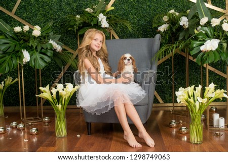 Little cute girl with blond hair in a white dress holding a small dog and white flowers, lilies and orchids on a green background