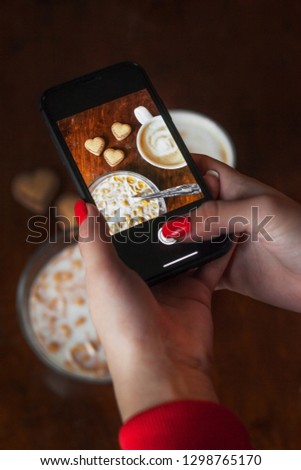 close-up of hands with red manicure with mobile phone taking pictures of coffee and cookies in the form of hearts on the table