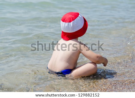 Little boy sitting in the shallows at the seaside cooling off in the refreshing water looking away from the camera