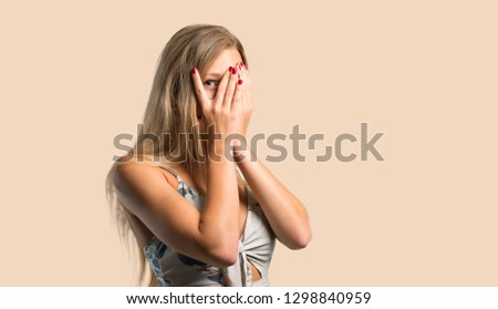 Young blonde woman covering eyes by hands and looking through the fingers on ocher background
