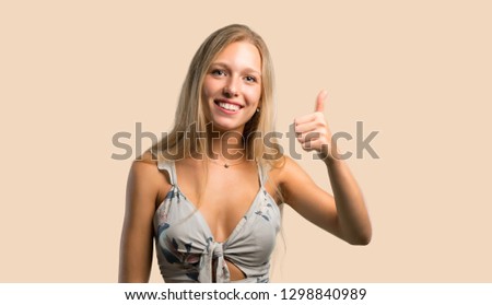 Young blonde woman giving a thumbs up gesture and smiling because something good has happened on ocher background
