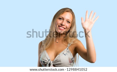 Young blonde woman saluting with hand with happy expression on blue background