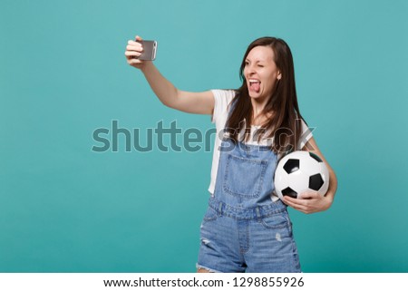 Funny young girl football fan with soccer ball doing selfie shot on mobile phone showing tongue isolated on blue turquoise background. People emotions sport family leisure concept. Mock up copy space