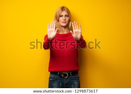 Blonde woman over yellow wall making stop gesture for disappointed with an opinion