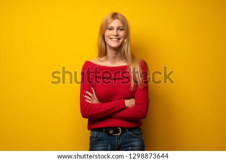 Blonde woman over yellow wall keeping the arms crossed in frontal position