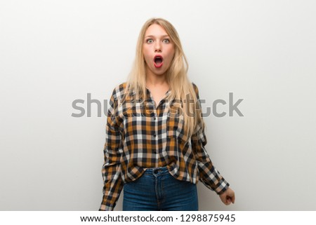 Blonde young girl over white wall with surprise and shocked facial expression