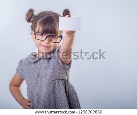 Cute smiling child with glasses holding discount white card in her hands. Kid with credit card. Little girl showing empty blank paper note, copy space. Isolated on blue background 