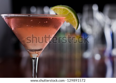 chilled cosmopolitan cocktail served in a  martini glass on a busy bar with a shallow depth of field with blurred glasses and lights in the back, garnished with a lime