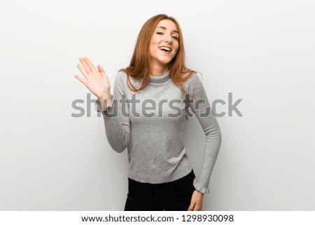 Redhead girl over white wall saluting with hand with happy expression