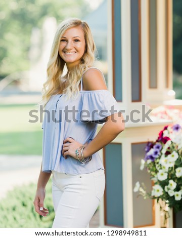 One attractive blonde hair caucasian high school senior posing for senior pictures. Female Teenager portrait standing outdoors at park in summer.