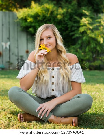One attractive blonde hair caucasian high school senior posing for senior pictures. Female Teenager portrait outdoors at park holding yellow flower in summer.