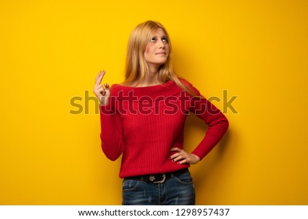 Blonde woman over yellow wall with fingers crossing and wishing the best