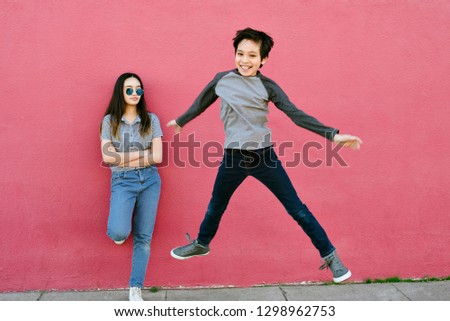 A teen sister looks onto her younger brother as he leaps into the air.  She is stoic and he is happy