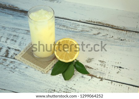 A glass cup with a refreshing juice and ice, on a textile stand and cut half of a lemon with leaves lies on a white wooden table. Close-up.
