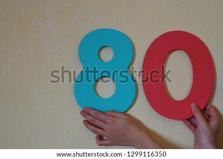 80 - hands holding colorful number eight and zero or eighty on mild yellow wall background with copy space for text. 80th anniversary or birthday design.
