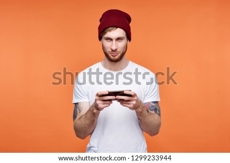 a man in a red cap holds a phone in his hands, a tattoo on his hand an orange background                             