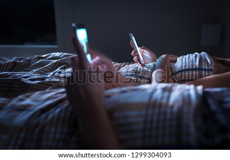 Bored distant couple ignoring each other lying in bed at night while using mobile phones. Smartphone addict. Obsessed and distracted man and woman texting. Addiction to social media and technology. Royalty-Free Stock Photo #1299304093