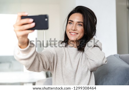 Photo of brunette woman 30s taking selfie on cell phone while sitting on couch in bright apartment
