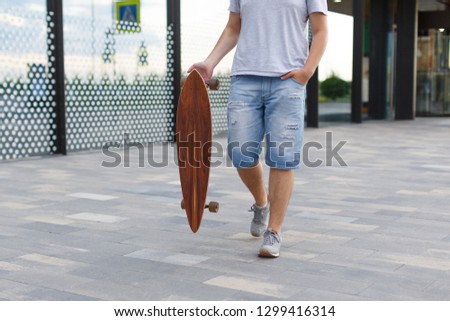 Close up street picture of man in denim shorts and grey sneakers walking with a longboard in right hand/ urban area, moving fast, hipster skateboarding in city/ people, lifestyle and sport concept.