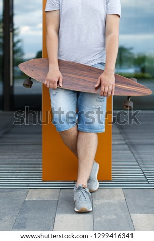 Vertical outdoor portrait of young man in denim shorts and grey sneakers holding longboard in hands against the glass case/streets of downtown district, urban area/people, lifestyle and sport concept