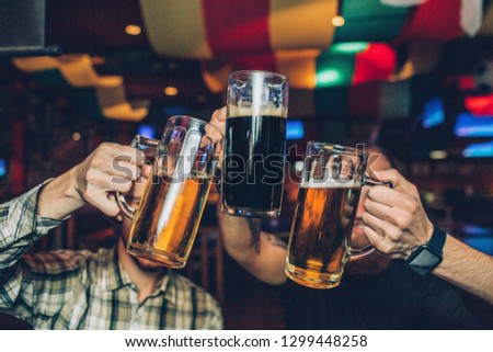 Picture of young men sitting in pub and holding mugs of dark and light beer together.