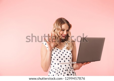 Beautiful young blonde woman wearing dress standing isolated over pink background, holding laptop computer, waving