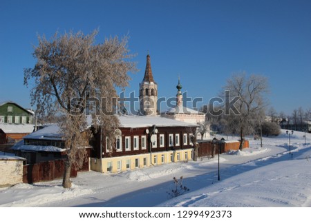 Suzdal white old church and belfry behind house roof on snowy street under blue sky