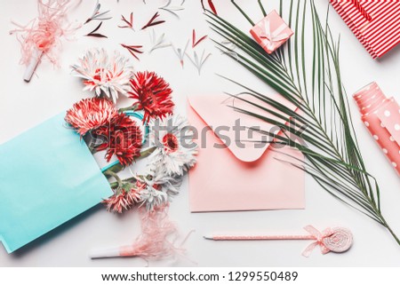 Female greeting objects flat lay in coral and turquoise colors. Shopping bag with Flowers bunch, blank envelope with pen, palm leaves and wrapping paper on white background, top view.