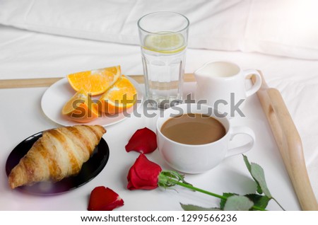 Romantic Valentines Day breakfast in bed, tray with croissant, cup of coffee espresso with milk, fresh oranges, glass of water with lemon and red rose. Good morning concept. Enjoy slow life.