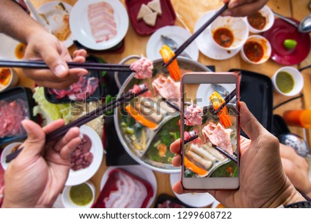 Hand holding a smartphone to use application where to eat buffet with blurred Shabu buffet background ,Freshly sliced pork meat with chopsticks over hot pot and vegetables for shabu and Japanese food.