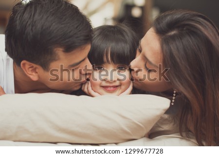 Young mother father and daughter enjoying with lie on the stomach in the bed, happy family smiling at home. mother and father kiss daughter at center