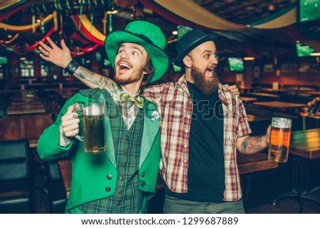 Happy young men holding mugs of beer and singing together in pub. They celebrate saitn patrick's day. Guy on left wear green suit