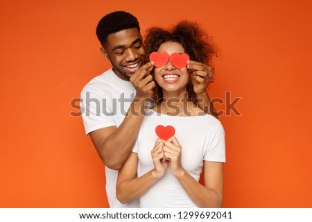 I love you. Happy young man is covering female eyes with surprise and giving her heart cards, orange studio background