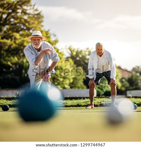 Elderly man playing boules in a playground with his playmate standing in the background. Old man in hat throwing a boules in a lawn with blurred boules in the foreground. Royalty-Free Stock Photo #1299744967
