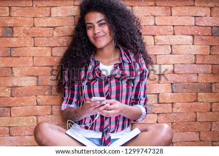 Happy student woman with afro hairstyle sitting with brick wall background using cell phone and smiling