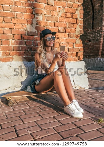 Girl in a baseball cap in the hands of the phone. Happy smiling sitting on a skateboard, longboard. In the summer in the city of the brick wall.