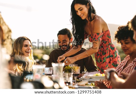 Beautiful woman serving food to friends sitting at dinner table. Group of friends having a party together outdoors.