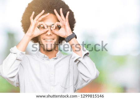 Young african american man with afro hair wearing glasses doing ok gesture like binoculars sticking tongue out, eyes looking through fingers. Crazy expression.