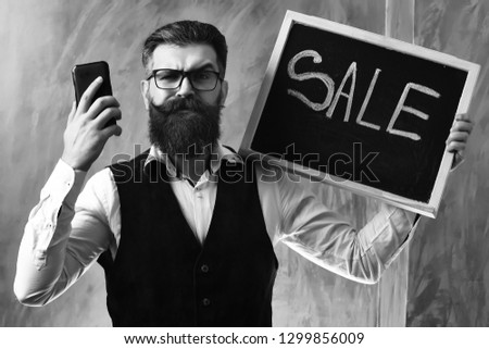 Bearded man, long beard. Brutal caucasian unshaven squinting hipster with glasses and moustache holding cell phone and sale inscription on blackboard on brown studio background