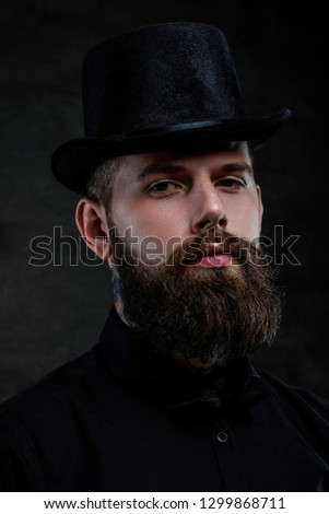 Close-up portrait of an old-fashioned bearded man with tattoos on his neck wearing a top hat, isolated on a dark background.
