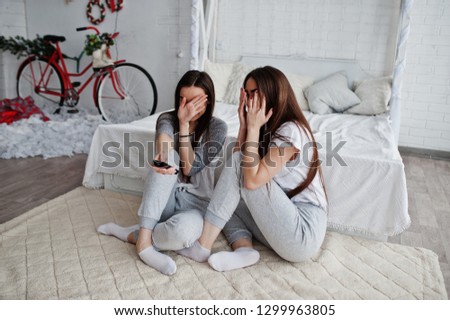Two twins models girls while click the remote control from the TV on studio and hiding faces, becouse they watched scary movie. They are frightened and shocked.